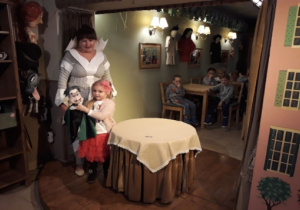 Aktorka na scenie stojąca przy stoliku oraz dziewczynka trzymająca pacynkę.